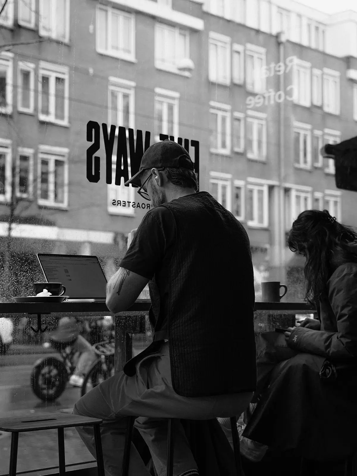 Black-and-white cafe scene with a person working on a laptop beside a large window, coffee cups on the counter, and another seated customer nearby.