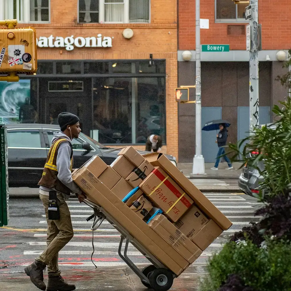 Delivery worker moving a loaded hand truck of cardboard packages along a city street.