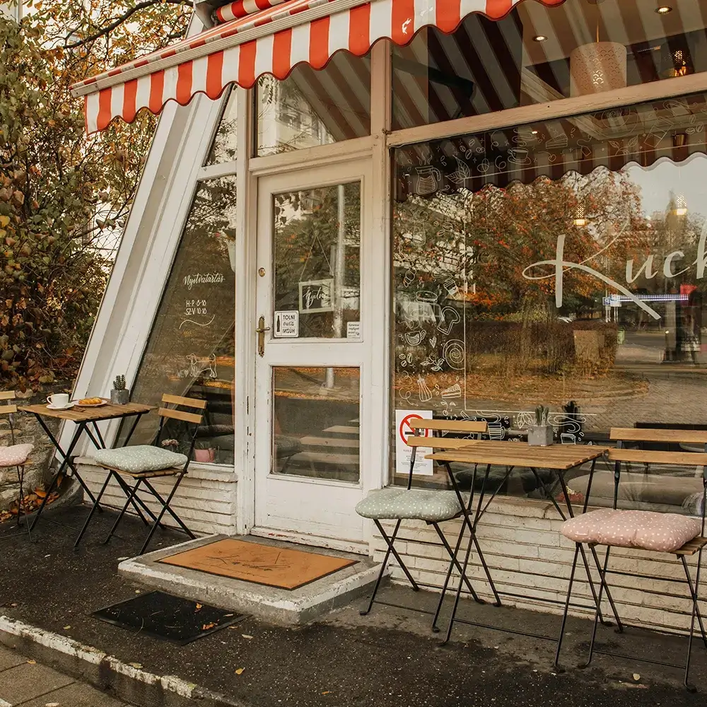 Local café storefront with striped awning, outdoor tables, large windows, and autumn trees.